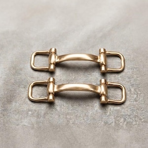 May include: Two identical gold-toned metal hardware connectors, each featuring a central curved bar flanked by two rectangular loops on either side. These metallic components have a smooth, polished finish and a symmetrical design.