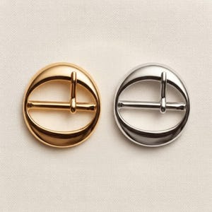 May include: Two circular belt buckles with a smooth, polished finish. One buckle is gold-toned, and the other is silver-toned. Each buckle features a central horizontal bar and a vertical prong, creating a simple, rounded frame design for fastening.