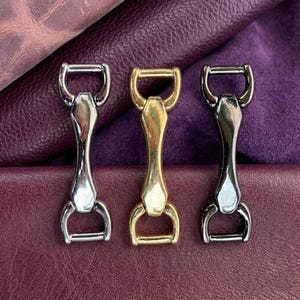 Può includere: Tre connettori metallici a forma di anello a D, con finiture argento, oro e canna di fucile. I connettori sono centrati con una barra curva e due anelli a D. Lo sfondo è in pelle strutturata, viola scuro e bordeaux.
