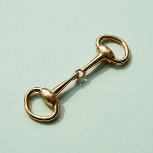 May include: A gold-coloured horse bit, a piece of equestrian equipment, is displayed against a light teal background. The bit features two rounded rings connected by a central bar with a small ring. The metal has a polished, reflective surface.