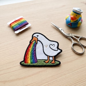 May include: An embroidered patch featuring a white goose vomiting a rainbow. The patch has a black border and is on a green patch of grass. Also pictured are a rainbow spool of thread, a rubber ducky in a sailor outfit, and small scissors.