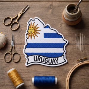 May include: An embroidered patch in the shape of Uruguay's map, featuring the country's flag and the word "URUGUAY". The patch is surrounded by sewing tools, including scissors, needles, thread, and an embroidery hoop. A gourd with a metal straw is also present.
