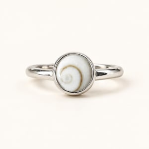 Könnte beinhalten: Ein silberfarbener Metallring mit einer kreisförmigen Fassung, die eine glatte, weiß-beige Einlage aus einer Muschel mit Spiralmuster enthält. Die Ringschiene ist poliert und massiv, mit einem schlichten, abgerundeten Design um das Zentrum.