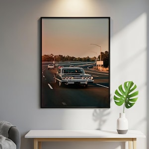 Könnte beinhalten: Ein gerahmter Wandkunstdruck, der ein schwarzes Oldtimer-Auto auf einer mehrspurigen Autobahn bei Sonnenuntergang zeigt. Die Szene zeigt die Rückansicht des Fahrzeugs mit markanten Rücklichtern, umgeben von anderen Autos unter einem orangefarbenen Himmel.