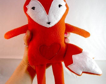 May include: A plush orange fox toy with white accents, a heart on its chest, and a white tail. The fox is being held by a hand.
