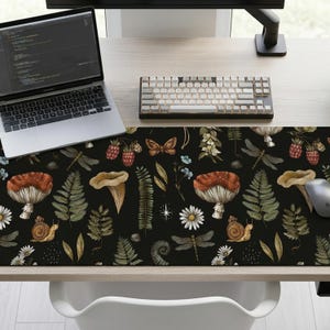 Op de afbeelding: Een zwarte bureaumat met een botanisch ontwerp met paddenstoelen, varens, libellen en bessen. De mat heeft een herhalend patroon van natuurlijke elementen in bruin, groen en wit. Een laptop, toetsenbord en muis liggen op het bureau.