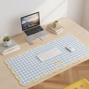 May include: A blue and white gingham desk mat with a scalloped yellow border. A white keyboard, mouse, and laptop are on the mat. A small succulent in a white pot, books, and a mug are also on the wooden desk.