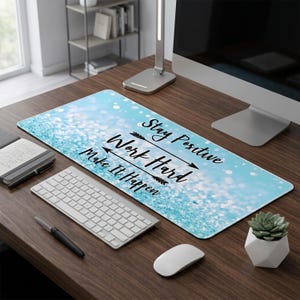 以下が含まれることがあります： ライトブルーのグリッターデザインと「Stay Positive, Work Hard, Make It Happen」の文字が黒でプリントされた長方形のデスクマット。白いキーボード、マウス、ペンが木製の机の上にあります。