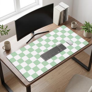 May include: A desk mat featuring a green and white chequered design. A black keyboard, white mouse, and a curved computer monitor are on the mat. The desk also has a mug, a small potted plant, and a stack of books.