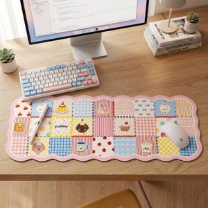May include: A patterned desk mat with a patchwork design. The mat features illustrations of cupcakes, cherries, and a puppy. It has a pink scalloped border and is paired with a white keyboard and mouse. The mat is designed for a desk.