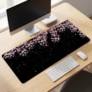 Op de afbeelding: Een rechthoekige bureaumat met een zwarte achtergrond en een trapsgewijs patroon van roze kersenbloesems en bloemblaadjes die vanaf de bovenrand naar beneden vallen. De mat ligt op een houten bureau onder een computermonitor, toetsenbord en muis.