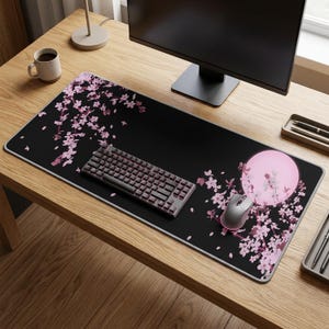May include: A black extended mouse pad with a pink floral design and a white border. A gray keyboard and mouse are on the pad. A computer monitor, a mug, and desk accessories are also visible.