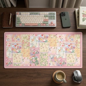May include: A floral patterned desk mat with a pink border, a keyboard, a phone, and a mouse on a wooden desk. The mat features a patchwork design with flowers, butterflies, and birds. The keyboard has pink and green keys.