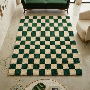 May include: A rectangular, plush rug with a checkerboard pattern of dark green and cream squares. The rug is placed on a light-colored floor, with a green sofa and cream-colored armchair in the background. The rug's design adds a modern touch to the room.