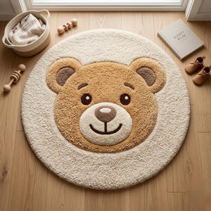Op de afbeelding: Een ronde, beige en bruine teddybeer-vloerkleed. Het kleed heeft een lachend berengezicht in het midden, met een lichtbeige rand. Houten speelgoed, een klein mandje, een boek met de titel "Sleepy Bear" en babyschoenen zijn ook zichtbaar.