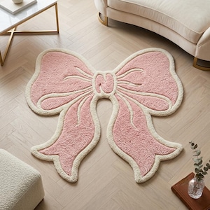 Può includere: Un morbido tappeto trapuntato a forma di grande fiocco, caratterizzato da un centro rosa chiaro testurizzato con un bordo spesso color crema che delinea i passanti e le estremità del design, creando un elemento decorativo per il pavimento.