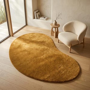 May include: An asymmetrical, kidney-shaped area rug in a solid mustard yellow colour with a plush, textured pile surface. The rug is positioned on a light-toned wooden floor, featuring a soft, dense appearance that covers a large floor space.