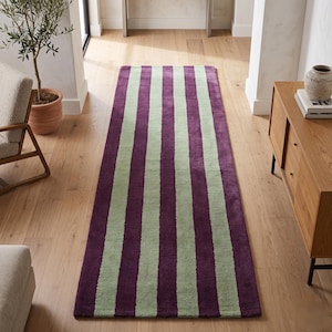 May include: A rectangular runner rug featuring a repeating vertical striped pattern in deep purple and soft sage green. The rug has a low-pile texture and is positioned lengthwise on a light-colored wooden floor, extending from the foreground.