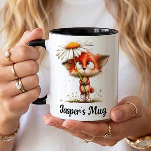 May include: A white ceramic mug with a black handle and rim, featuring a graphic illustration of a small orange fox wearing boots and holding a large white daisy. The text Jasper's Mug is printed in a black cursive font below the fox illustration.