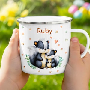 Puede incluir: Una taza de esmalte blanco con borde plateado que presenta un gráfico de dos mofetas, hojas verdes y pequeños corazones naranjas. El nombre Ruby está impreso en letras marrones sobre las mofetas. La taza tiene un asa lateral y acabado liso.