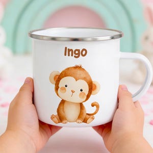 Può includere: Una tazza in smalto bianco con bordo argentato e manico, caratterizzata da un'illustrazione di una scimmia marrone e dal nome Ingo stampato in marrone sopra di essa. La tazza è tenuta da due mani, mostrando chiaramente il design frontale.