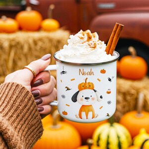 Op de afbeelding: Een witte emaille mok met een cartoon puppy met een pompoenhoed, de naam Noah en kleine Halloween-iconen zoals spinnen en snoepjes. De mok is gevuld met slagroom en kaneelstokjes voor een feestelijke, seizoensgebonden uitstraling.
