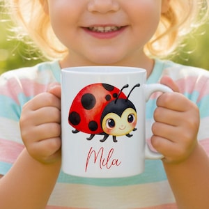 Może przedstawiać: Biały ceramiczny kubek z kolorową ilustracją biedronki z czerwonym pancerzem i czarnymi kropkami. Pod biedronką znajduje się napis Mila wykonany czerwoną kursywą. Kubek posiada zakrzywione ucho po prawej stronie ułatwiające trzymanie naczynia.