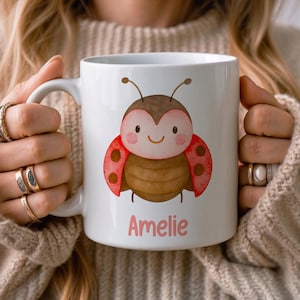 Op de afbeelding: Een witte keramische mok met een aquarelillustratie van een bruin en rood lieveheersbeestje met een glimlachend gezicht. Onder het lieveheersbeestje staat de naam Amelie gedrukt in een zachtroze, afgerond lettertype.
