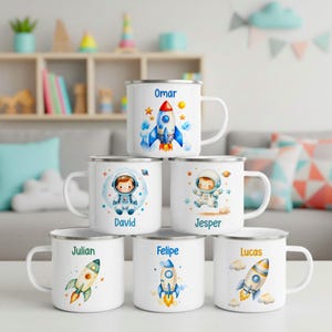 Puede incluir: Una pila de seis tazas de esmalte blanco con coloridas ilustraciones de temática espacial de cohetes y astronautas. Cada taza está personalizada con un nombre: Omar, David, Jesper, Julian, Felipe o Lucas, impreso en texto azul o naranja.