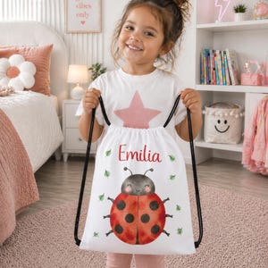 Peut inclure: Un sac à dos à cordon blanc orné d'une illustration de coccinelle rouge avec des points noirs, des feuilles vertes et le prénom Emilia écrit en rouge. Le sac possède des cordons noirs et est fabriqué dans un tissu léger.