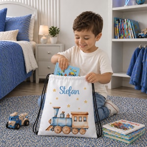 May include: A white drawstring backpack featuring a watercolour-style blue train illustration, yellow stars, and the name Stefan printed in blue script. The bag has black cord straps and is made of a smooth, lightweight fabric material.