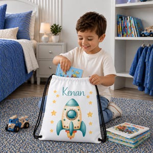 Op de afbeelding: Een witte rugzak met trekkoordsluiting, voorzien van een illustratie van een blauwgroene en beige raket, gele sterren en de naam Kenan in blauwgroen schrift. De tas heeft zwarte koordhengsels en is gemaakt van lichtgewicht, matte stof.