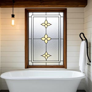 May include: A bathroom scene featuring a white, freestanding bathtub beneath a window with a stained glass design. The window has a wooden frame and a frosted glass panel with yellow floral accents. A black towel rack holds a white towel.