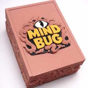 May include: A dusty rose-coloured box with the words "MIND BUG" in yellow, raised lettering. The box lid features a detailed relief of an eye, tentacles, and the text. The sides of the box have a repeating swirl pattern.