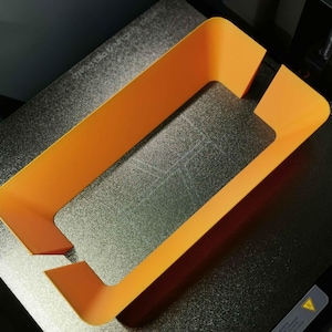 Könnte beinhalten: Ein orangefarbenes, rechteckiges 3D-gedrucktes Objekt mit einem ausgeschnittenen Bereich, das auf einer strukturierten, dunklen Oberfläche liegt. Die Form des Objekts deutet darauf hin, dass es sich um einen Rahmen oder eine Komponente für einen größeren Gegenstand handeln könnte.