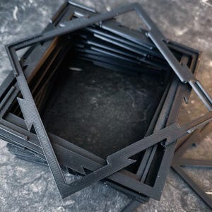 May include: A stack of black square frames with a geometric star-like cutout pattern. Each frame features thin, angular lines forming a central square opening with pointed extensions at the corners, creating a layered, textured appearance.