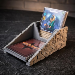 Può includere: Un portacarte dalla texture in pietra grigia con design inclinato a due scomparti. Una sezione contiene un mazzo di carte con retro rosso etichettato CLANK!, mentre l'altra sostiene una carta verticale intitolata Swap Meet con un'illustrazione di sfinge.
