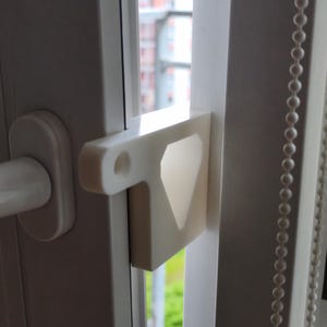May include: A white plastic window lock fitted to a white window frame. The lock features a geometric design with a diamond-shaped aperture. The window is ajar, revealing greenery and a building in the background.