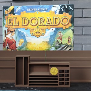 Può includere: Un organizer per giochi da tavolo in plastica marrone con scomparti rettangolari multipli e fessure per carte e gettoni. Un emblema circolare giallo è fissato sulla parte anteriore. L'organizer è posizionato davanti a una scatola di gioco El Dorado.