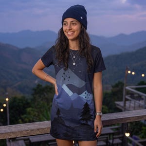 May include: A dark blue short-sleeved t-shirt dress featuring a graphic print of layered mountain silhouettes, pine trees, and a starry night sky. The dress is paired with a matching dark blue ribbed knit beanie with a small white star emblem.