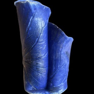May include: A cobalt blue ceramic vase with a leaf design. The vase has a unique shape, with two sections that resemble rolled leaves. The surface has a textured appearance, with visible veins mimicking the natural details of a leaf.