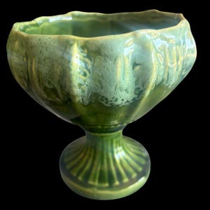 May include: A green ceramic pedestal bowl with a fluted base and a ruffled rim. The bowl has a glossy finish with variations in the green glaze, creating a marbled effect. The bowl is designed to hold fruit or other decorative items.