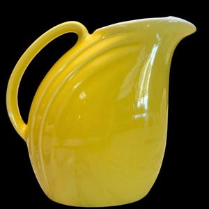 May include: A bright yellow ceramic pitcher with a curved handle and a spout. The pitcher has a glossy finish and a smooth, rounded body. The handle is arched and attached to the side of the pitcher. The background is black.