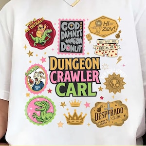 Op de afbeelding: Wit t-shirt met kleurrijke grafische ontwerpen. Het shirt heeft verschillende cartoon illustraties en tekst, waaronder "Dungeon Crawler Carl", "God Damnit Donut" en "Desperado Club". Het ontwerp bevat sterren, kronen en een dinosaurus.