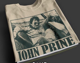 Op de afbeelding: Zandkleurig T-shirt met een zwart-wit afbeelding van een man met een zonnebril en de tekst "JOHN PRINE" eronder. Het woord "SAND" staat onderaan gedrukt. Het shirt is opgevouwen en gestapeld met andere shirts.
