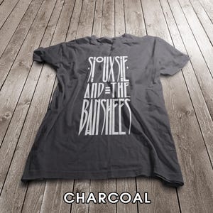 Puede incluir: Una camiseta de manga corta color gris carbón con texto gráfico blanco impreso en la parte delantera que dice Siouxsie and the Banshees en una fuente estilizada y alargada. La prenda tiene cuello redondo y un ajuste estándar con pliegues.