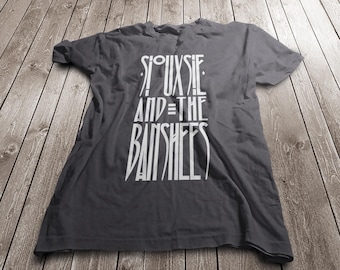 May include: A charcoal gray short-sleeved t-shirt featuring white graphic text printed on the front that reads Siouxsie and the Banshees in a stylized, elongated font. The garment has a crew neckline and a standard fit with visible fabric folds.