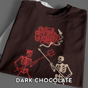 Könnte beinhalten: Ein dunkel schokoladenbraunes T-Shirt mit einer roten und cremefarbenen Grafik von zwei Skeletten, die Speere halten. Über den Skeletten steht der Text Sturgill Simpson in einer stilisierten roten Schrift mit einer kleinen Rakete.