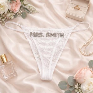 May include: White lace thong underwear with the text "MRS. SMITH" in silver rhinestones. The lingerie is displayed on a satin surface, with a ring box, perfume bottle, and flowers in the background.