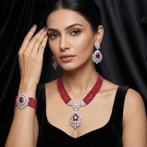 Op de afbeelding: Een vrouw draagt een robijn- en diamanten sieradenset. De set bevat een ketting met meerdere strengen met een hanger, bijpassende oorbellen en een armband. De sieraden hebben een combinatie van rode edelstenen en heldere diamanten, gezet in een zilveren of witgouden zetting.